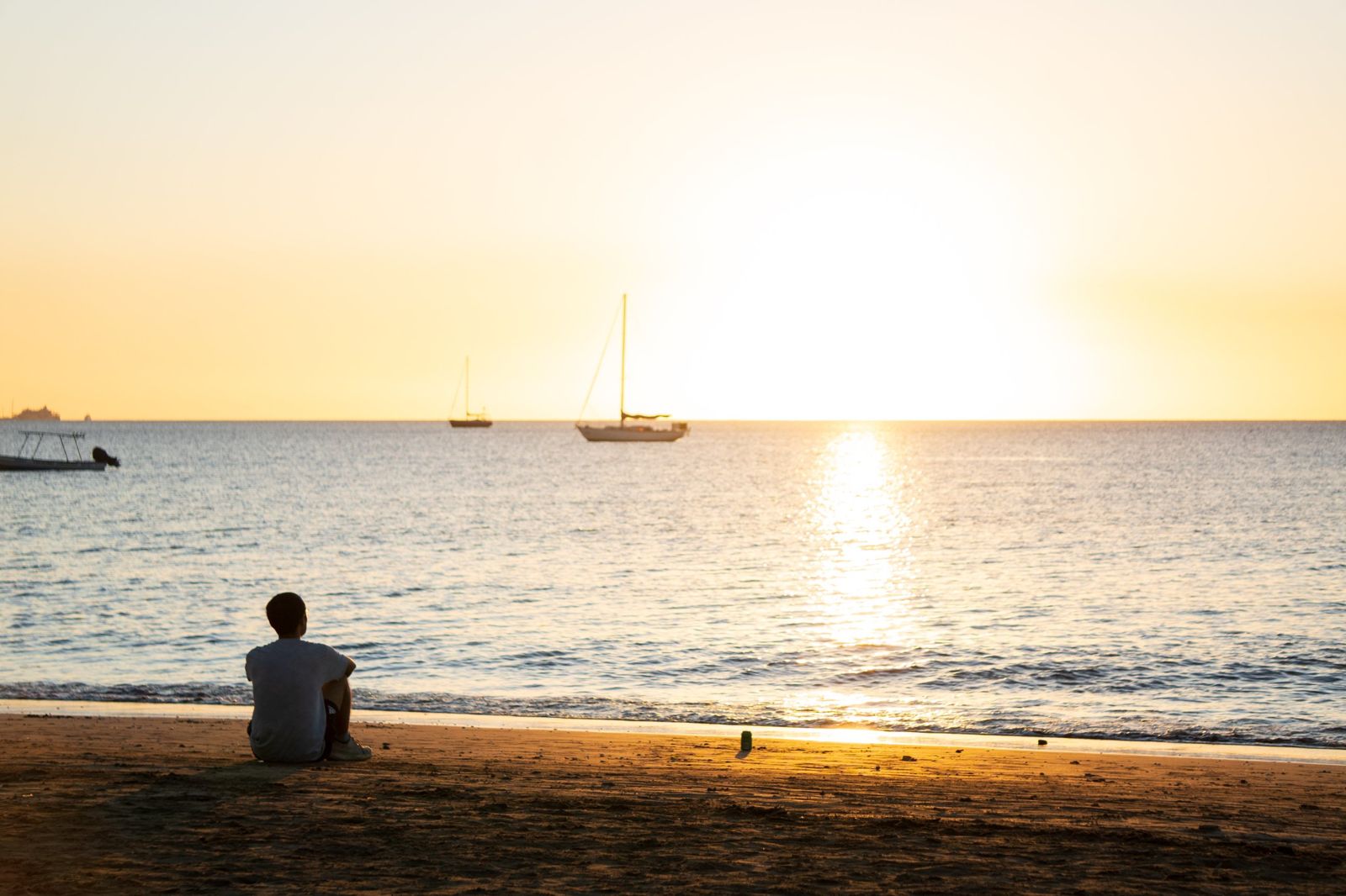 Man sitting on the sand watching sailboats at sunset