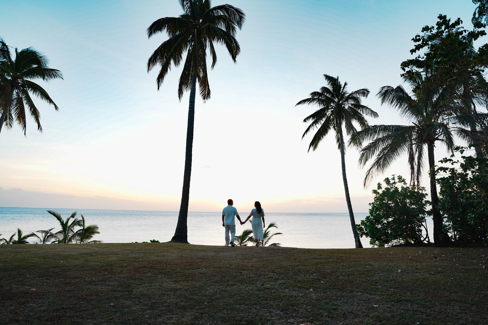 Couple in white holding hands under palms as the sun sets over the sea