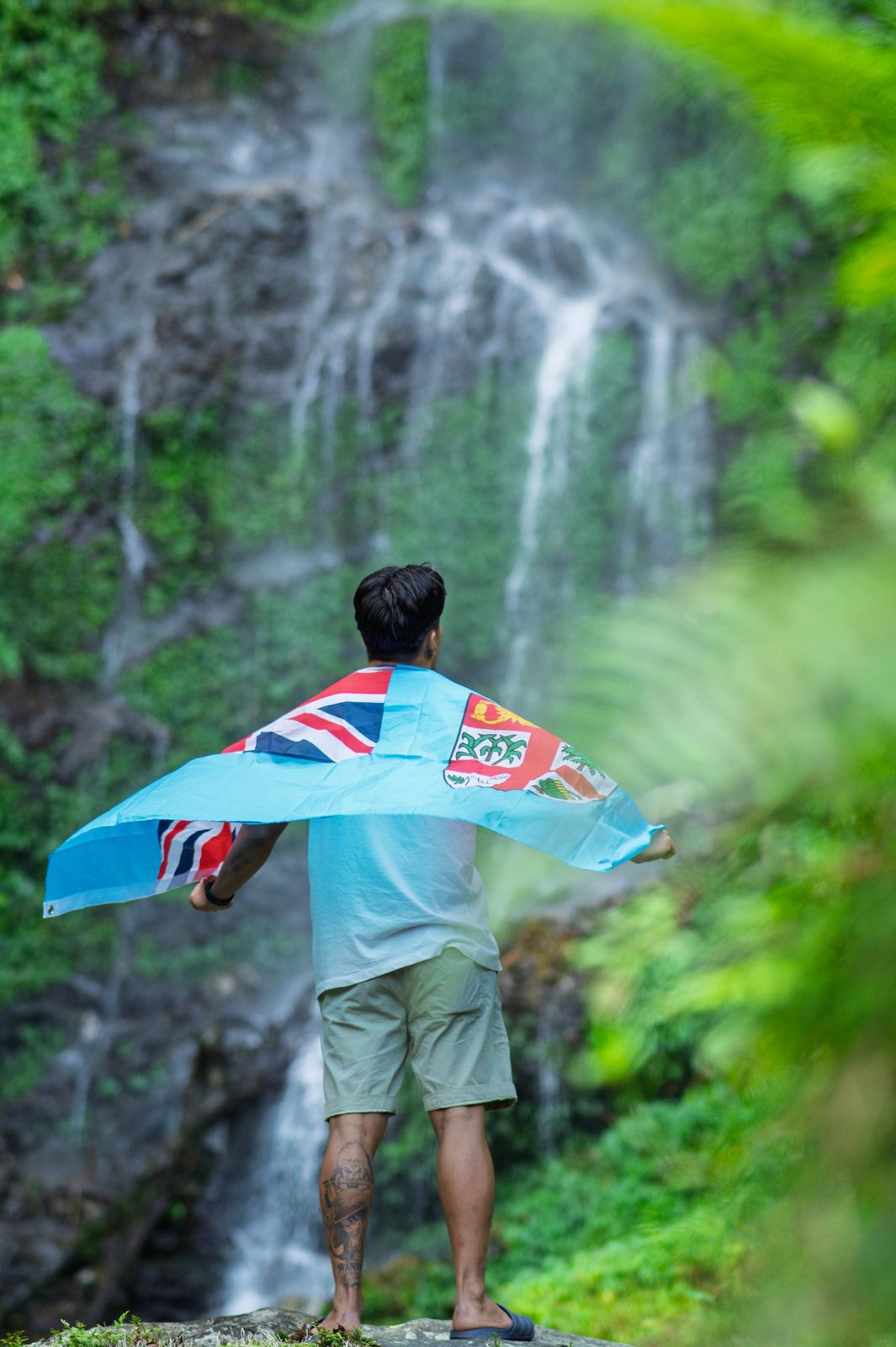 Man holding the Fiji flag across his back in front of a waterfall
