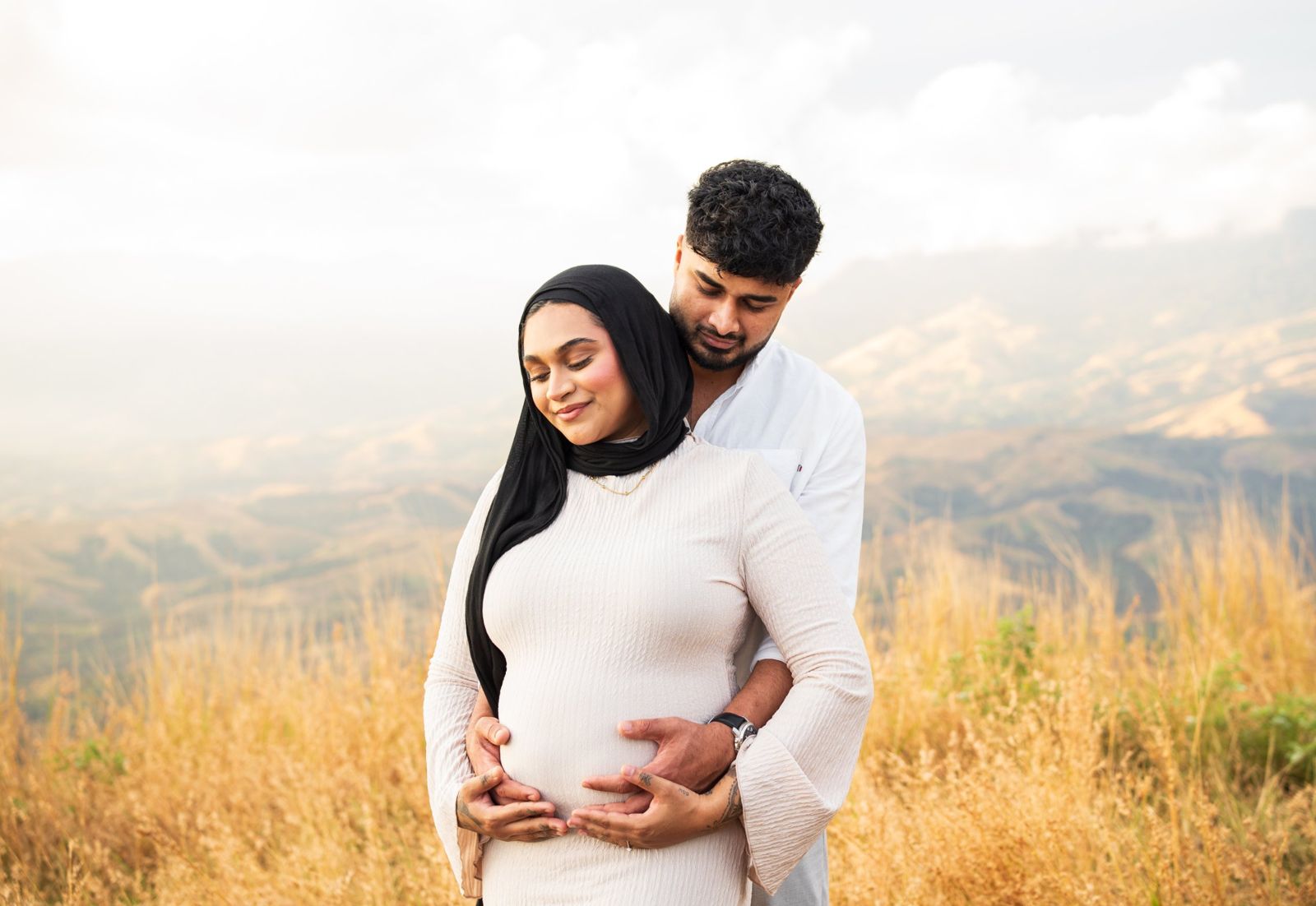 Expecting couple embracing in golden grass with hills behind